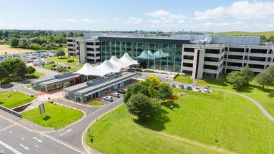 A birds eye view of a large, glass-fronted office building, surrounded by bright green grass on a sunny day.